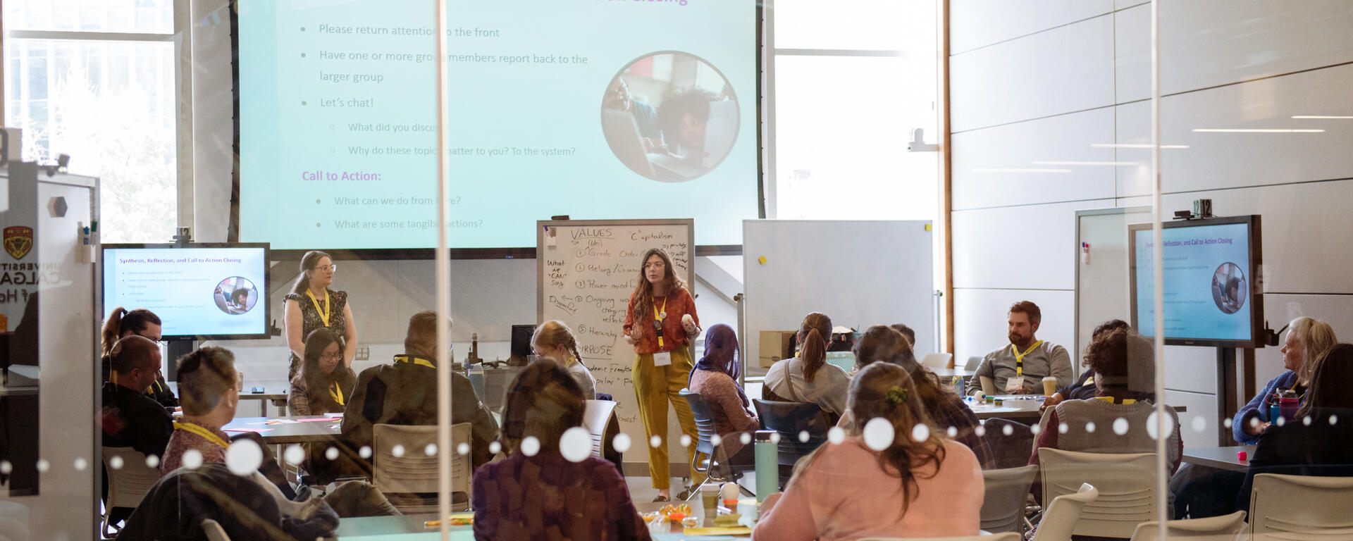 Looking inside a classroom with large windows and a speaker talking to students in front of a presentation.