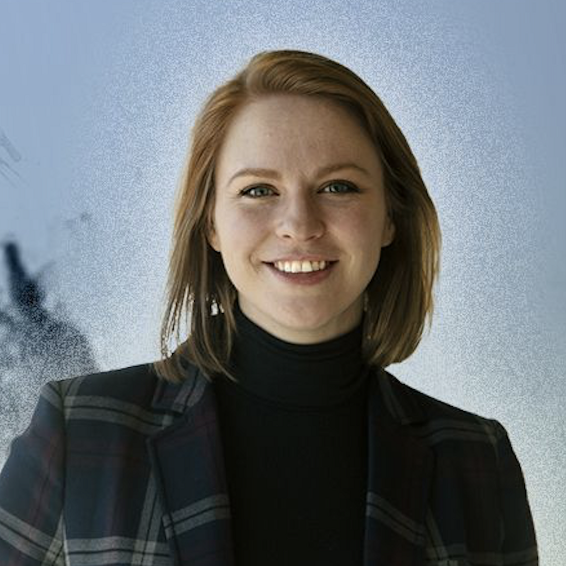 Stacey Copeland smiles in front of a blue background and is wearing a plaid blazer.