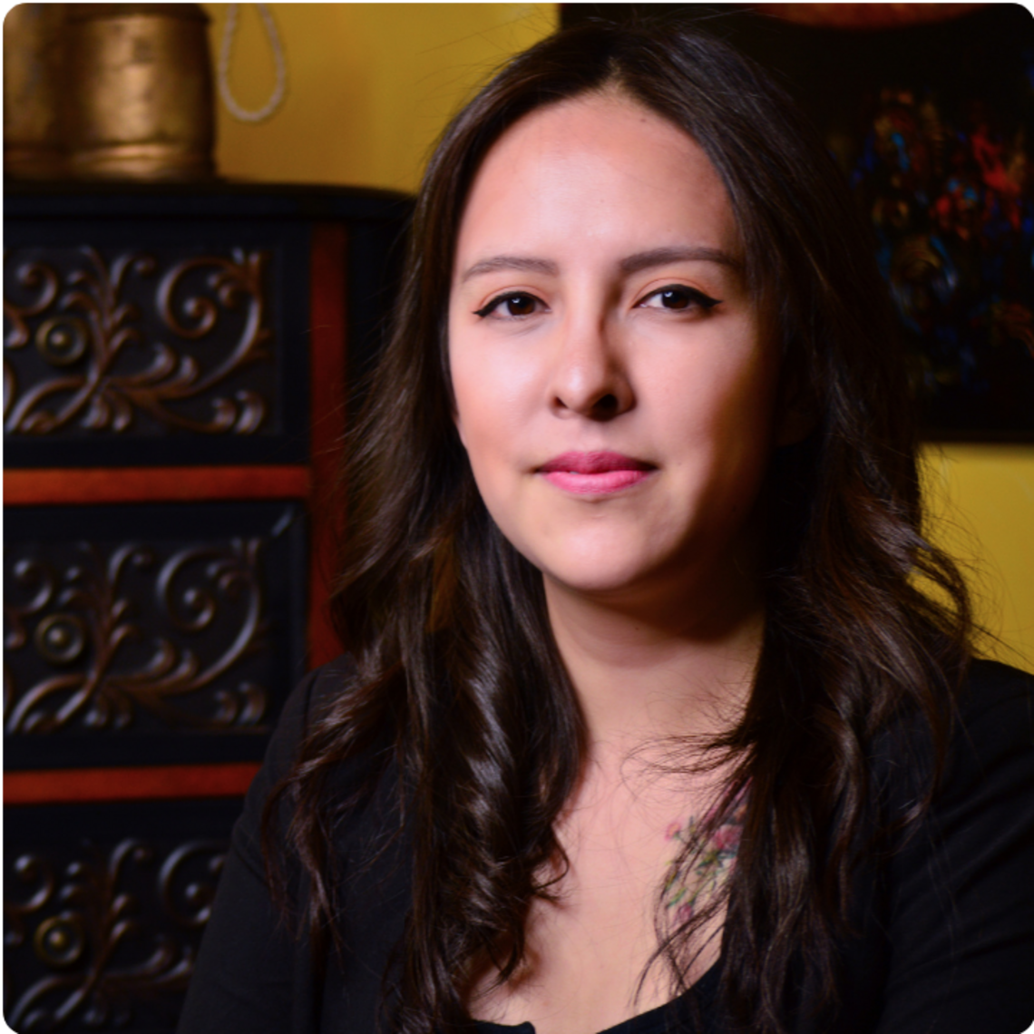Bernice Cancino, a woman with light skin and long dark hair, is inside beside an ornate piece of furniture looking ahead.