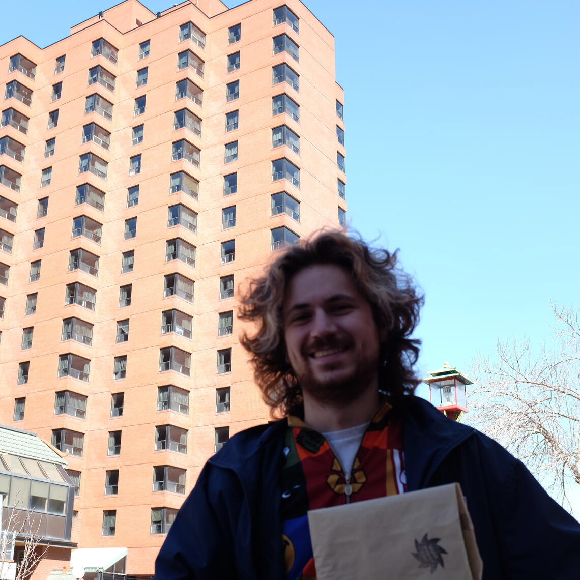 Brennan Chaudhry, a young man with fair skin and curly hair, is standing outside with city buildings and blue sky behind him.
