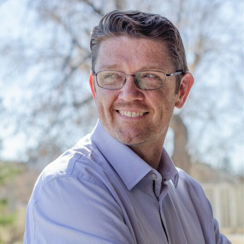 Blake Evernden, a white man with glasses, smiles outside and looks away.