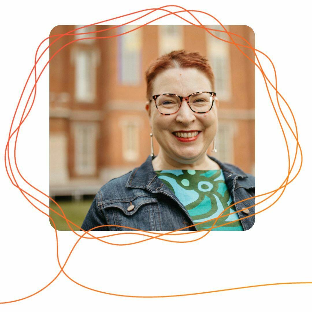 Cate Denial, a white woman with red hair and glasses, smiles outside with a brick building in the background.