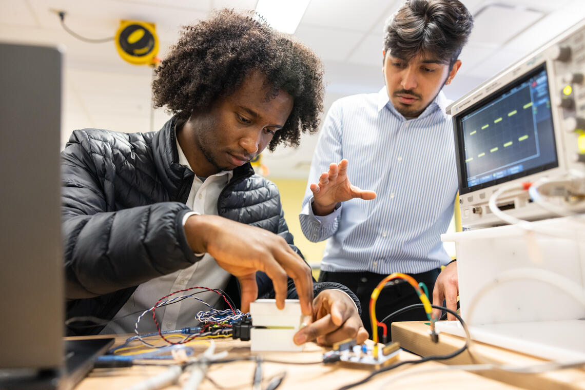 A Black student working with electronics with a brown teacher standing behind explaining.