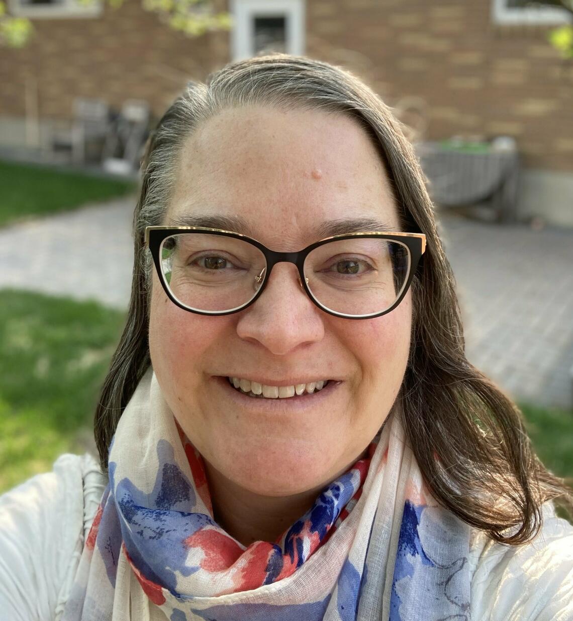 Nicole Sandblom, a fair-skinned woman with brown hair and glasses, is wearing a scarf outside and smiling.