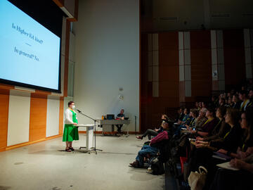 A presenter stands in front of a full auditorium of attendees