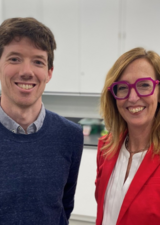 Martin MacInnis, a white man with light brown hair and wearing a blue cardigan over a collared shirt, stands beside Cari Din, a white woman with medium length blonde hair, wearing a red blazer and big red glasses. They are in a lab.