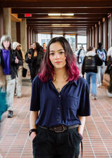 Emilie Lui, a young student with dark hair that has pink on the ends, stands in a busy hallway with blurred people walking around her. They are facing forward and have their hands in their pockets.