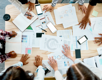 An overhead photo of people at a table with phones, notebooks and laptops on its surface